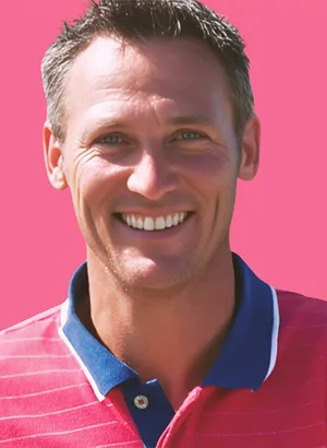 Homme souriant en polo rose avec col bleu sur fond rose.