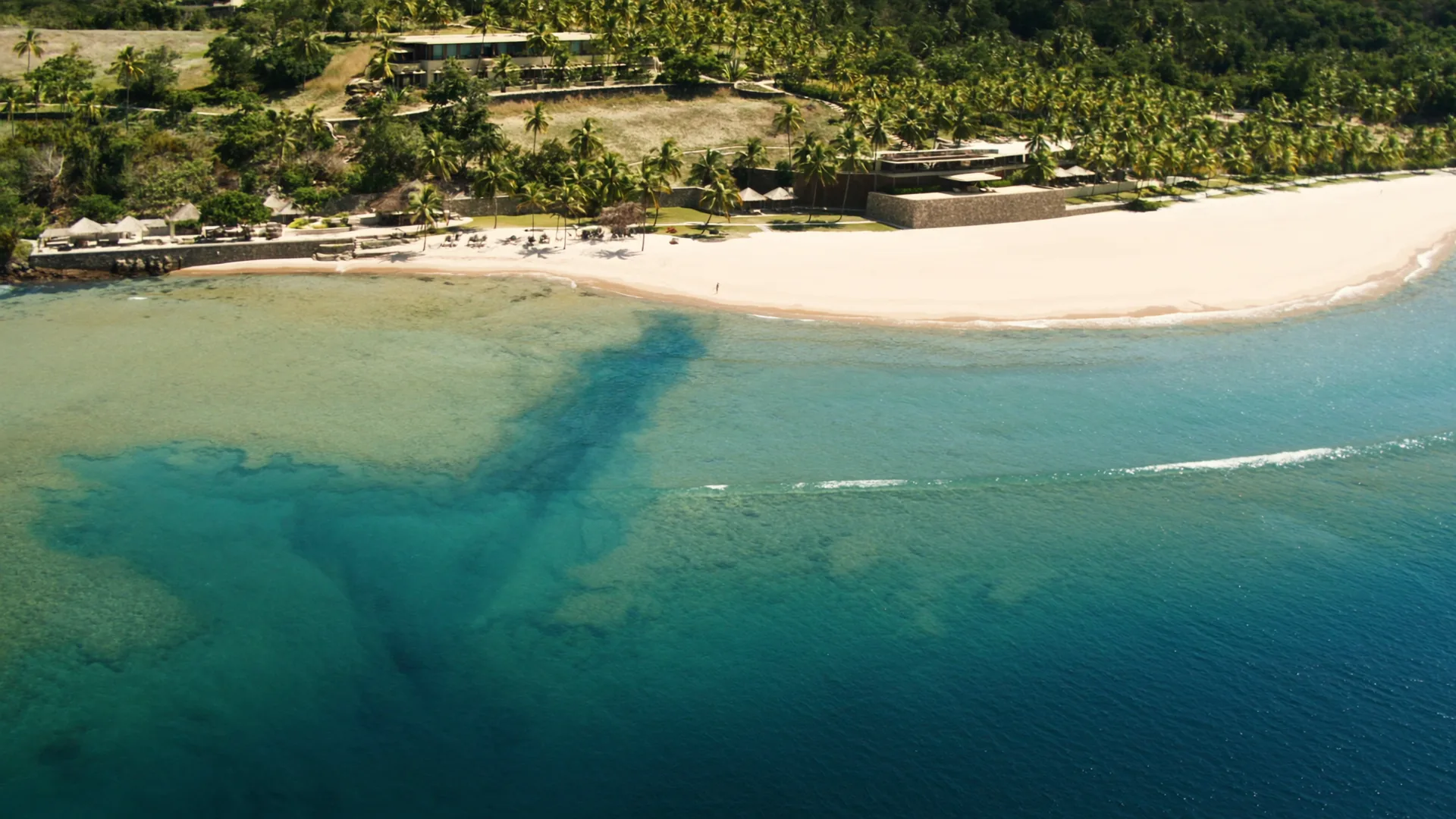 Aerial view of a luxury beachfront resort at golden hour, white sand meeting turquoise water, palm canopy framing low-rise architecture