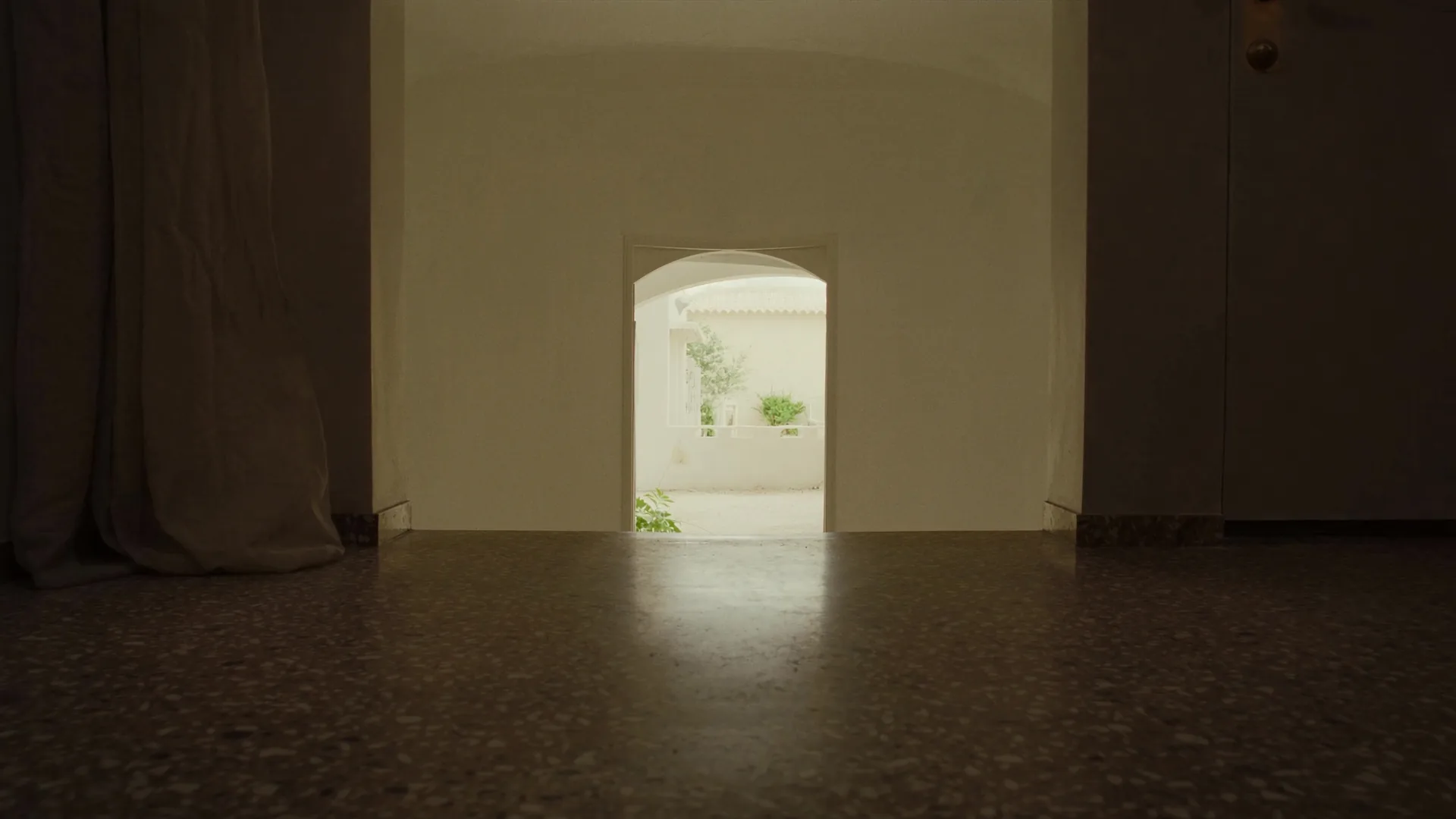 Minimalist hotel corridor with polished terrazzo floor, single arched doorway framing a Mediterranean courtyard, natural light and muted earth tones