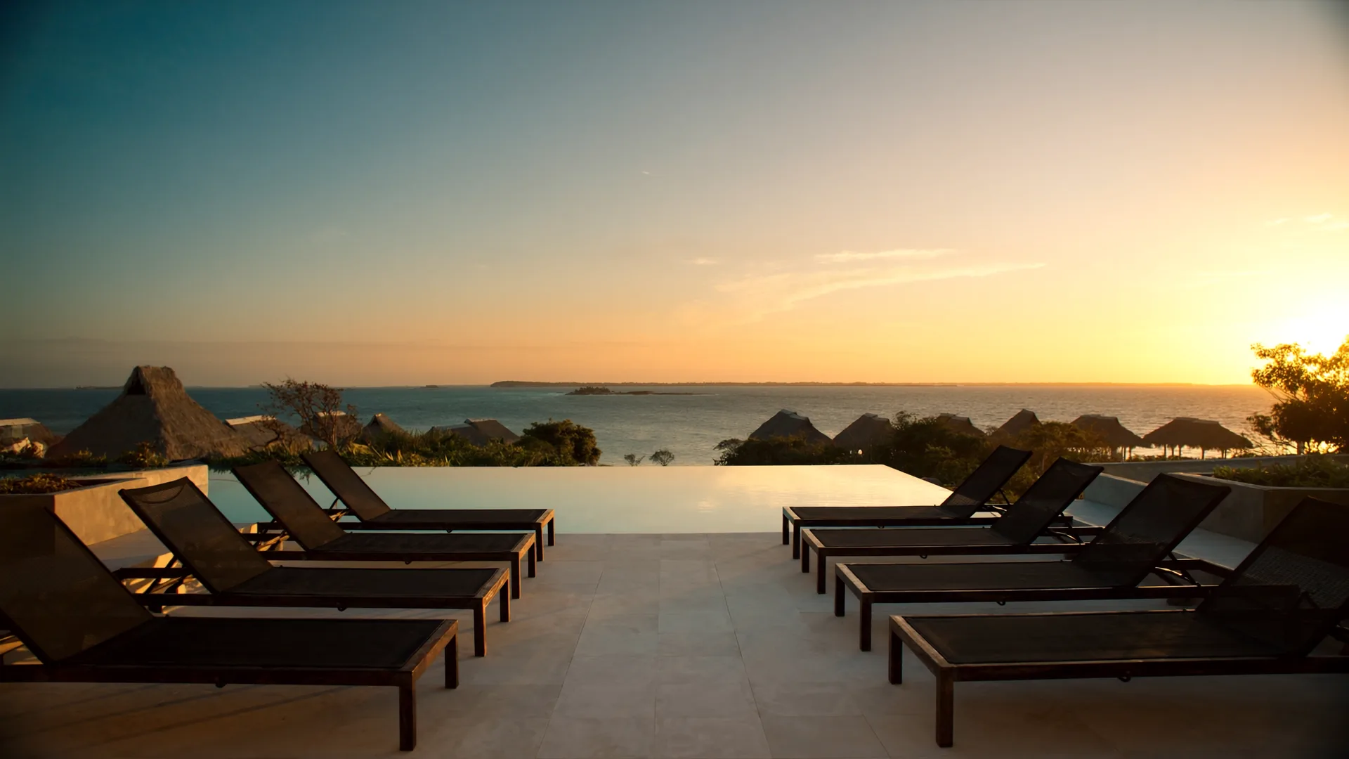 Resort infinity pool deck at dawn meeting calm Caribbean waters, teak loungers in geometric rows, tropical landscaping in warm golden light