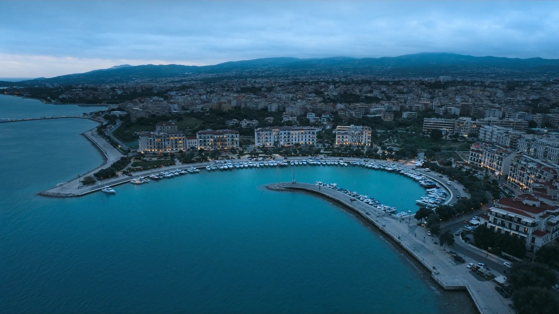 Aerial view of a Mediterranean harbour town at blue hour with several illuminated hotel facades along the waterfront